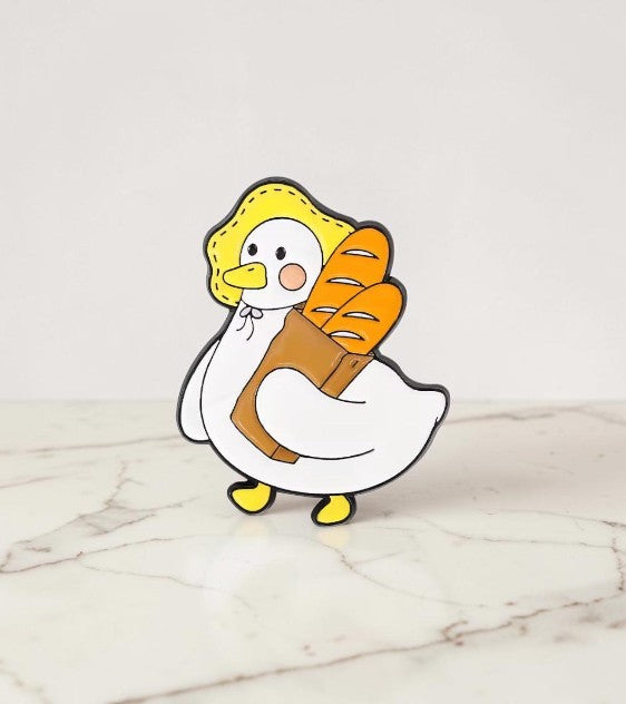 Pins & Co. enamel pin featuring a cute white duck wearing a yellow bonnet and carrying a paper bag of fresh baguettes.