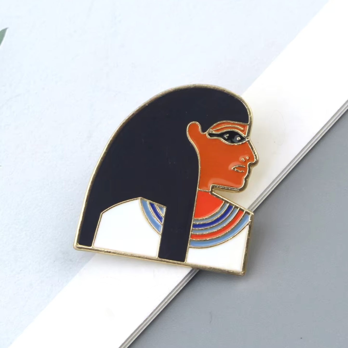 Pins & Co. Egyptian Queen enamel pin with gold-tone metal, black hair, and vibrant collar detailing on a white surface.