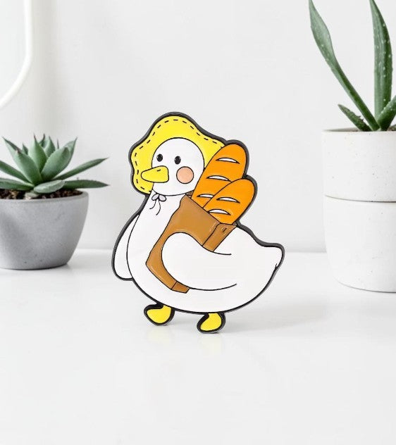 Pins & Co. enamel pin featuring a cute white duck wearing a yellow bonnet and carrying a brown bag of fresh baguettes.